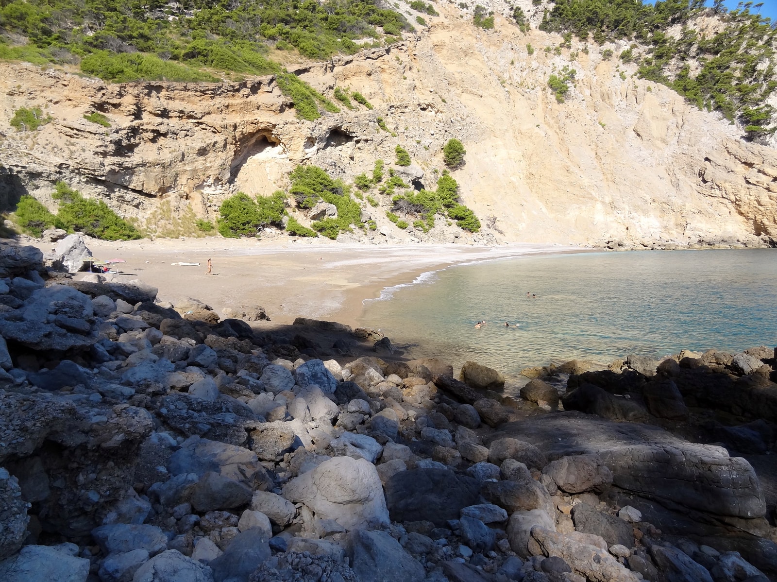 Rocky foreground frames a view of a remote pebble beach with clear water, enclosed by steep, rugged cliffs.