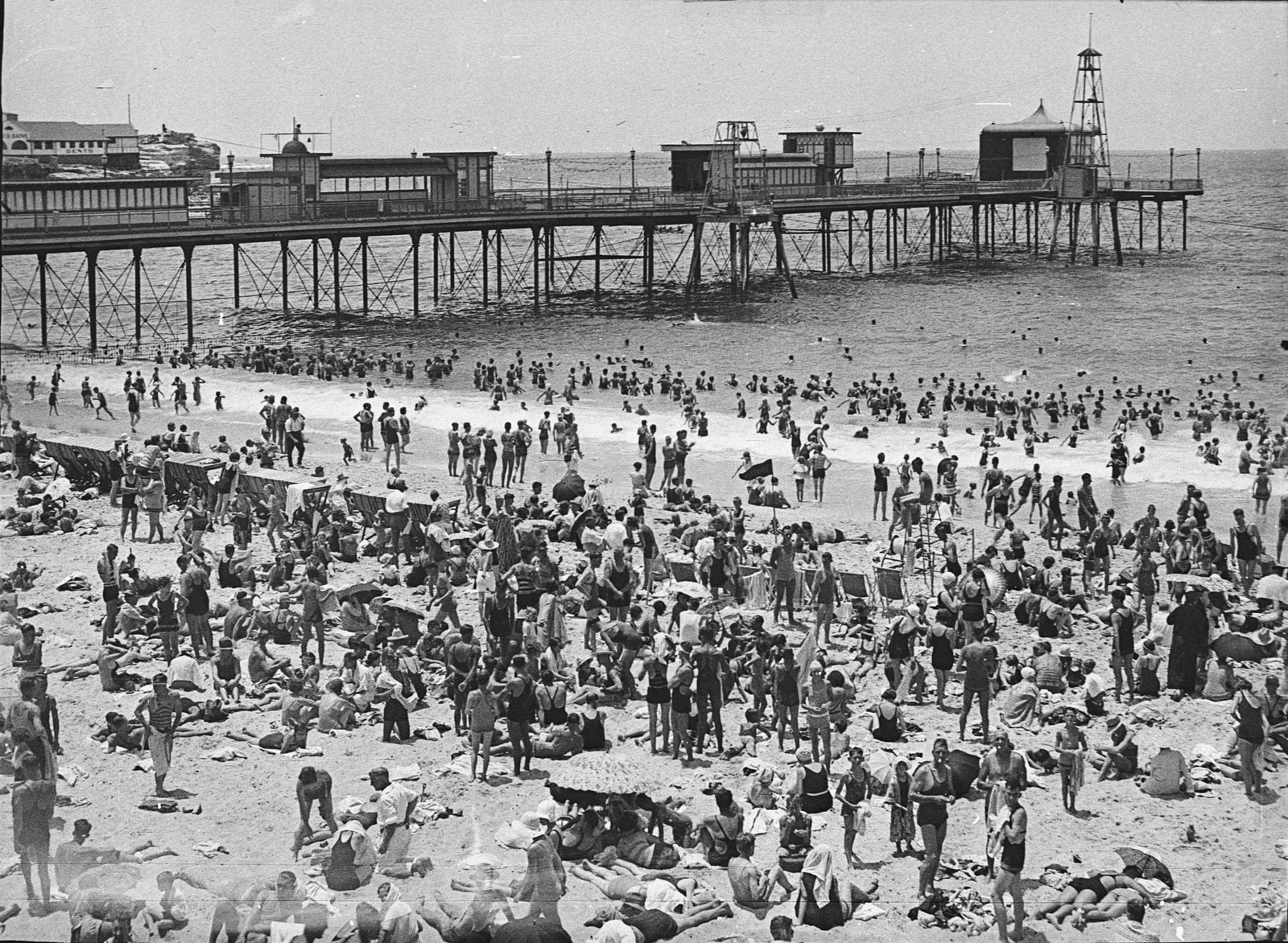 A black-and-white historical photo from 1928 shows large crowds on the beach under the old Coogee Pier.