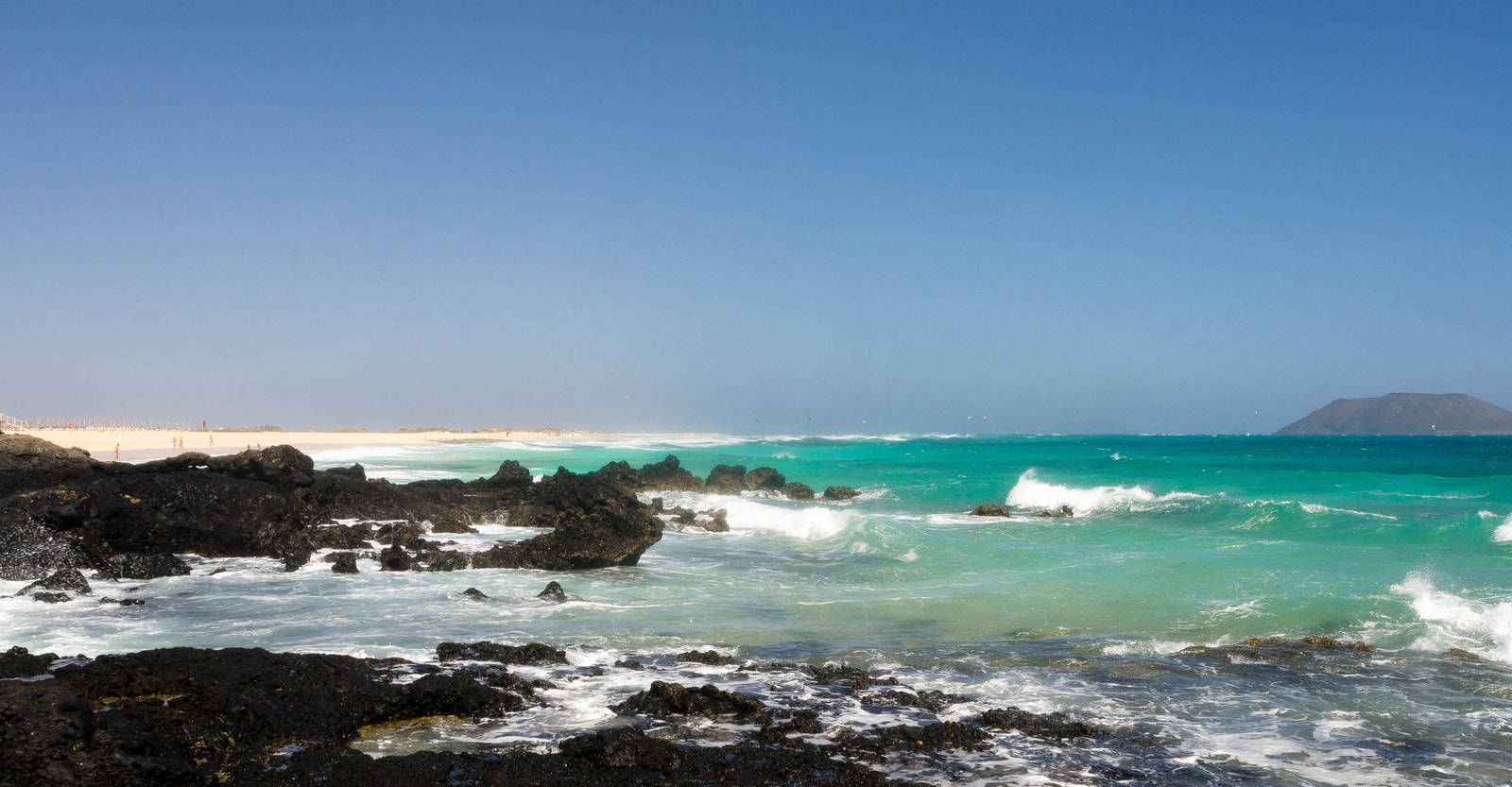 A wide white sand beach with dark volcanic rocks in the foreground, with turquoise water and gentle surf.