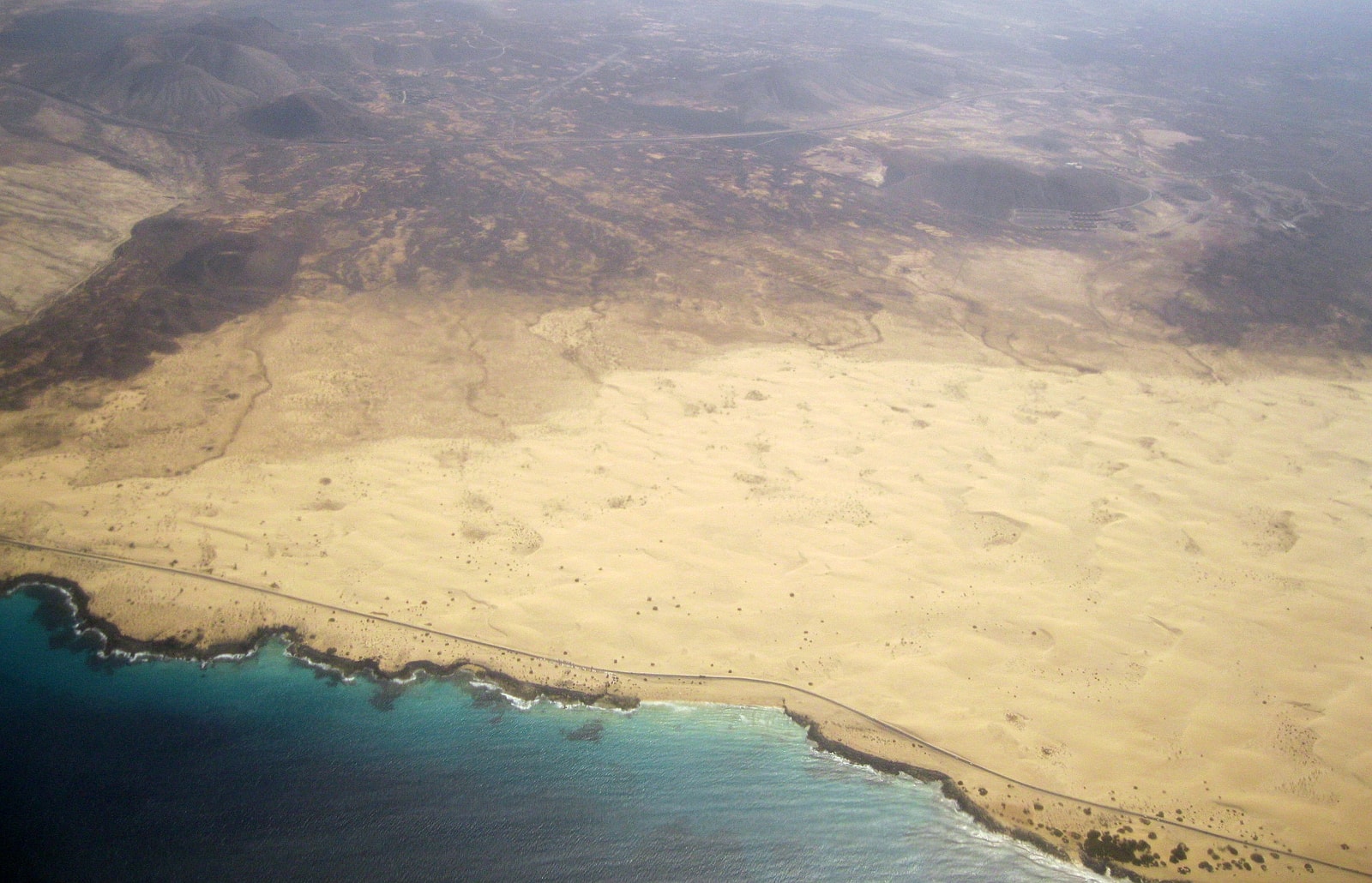 An aerial view shows the vast expanse of pale sand dunes in Corralejo Natural Park meeting the blue ocean.