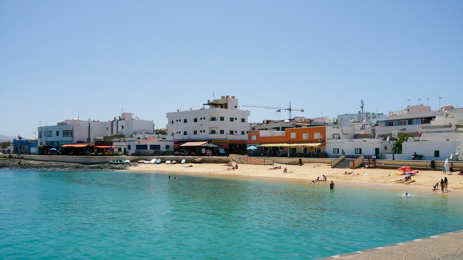A small, sheltered beach in Corralejo is lined with waterfront buildings and dotted with sunbathers and boats.