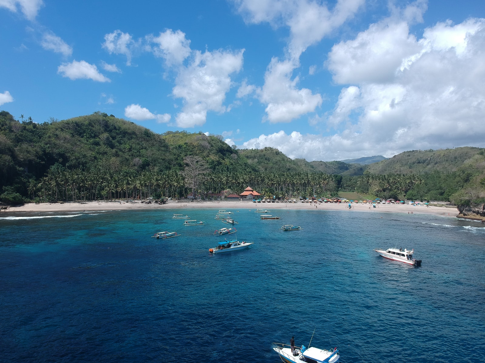A view of Crystal Bay on Nusa Penida, with several boats moored in the calm water, a sandy beach, and green hills in the background.