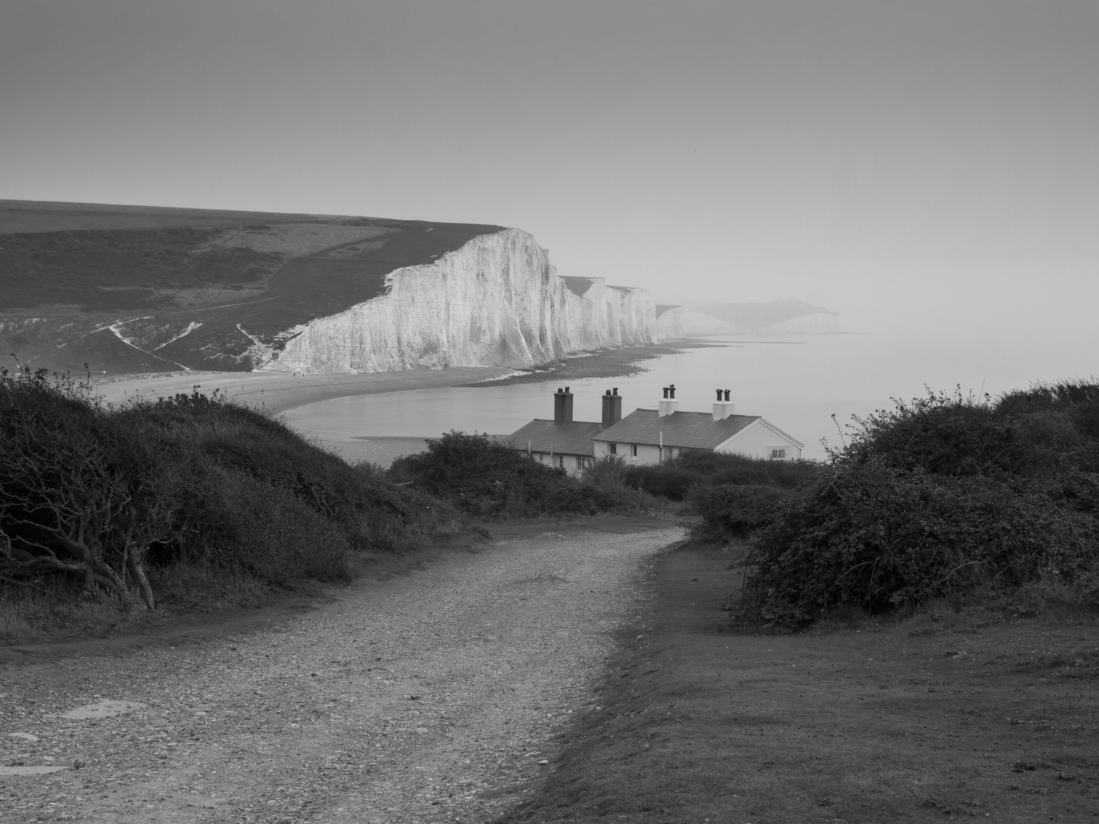 A black and white photo shows a path leading towards the Coastguard Cottages, with the dramatic Seven Sisters cliffs in the background.
