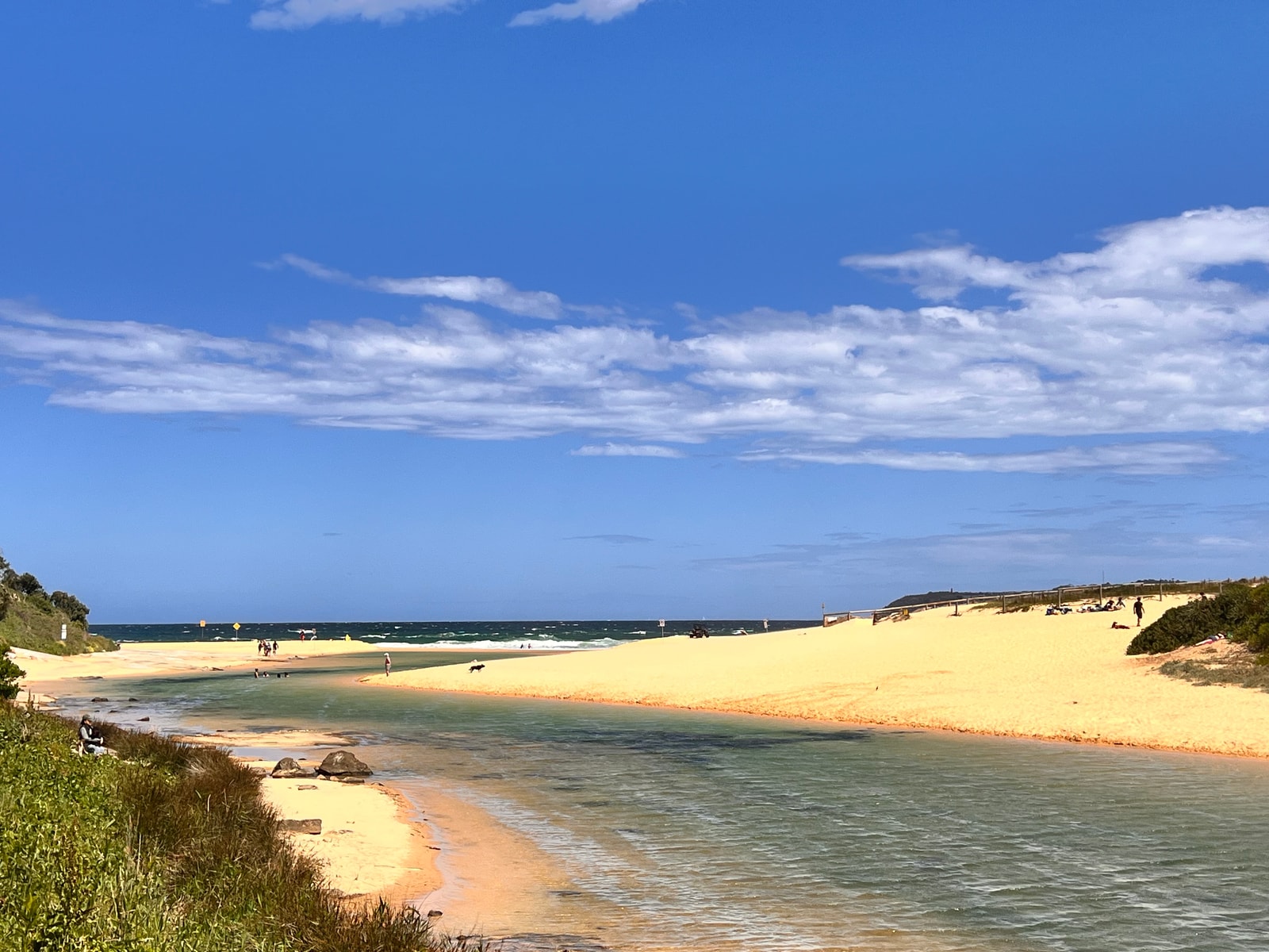 The serene Curl Curl Lagoon is pictured with its calm water reflecting the sky, flowing towards the ocean in the distance.
