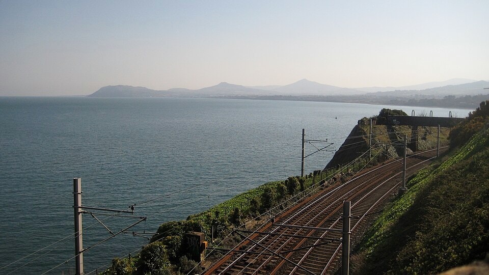 Train tracks run alongside the rocky coastline, with the blue sea visible just beyond.