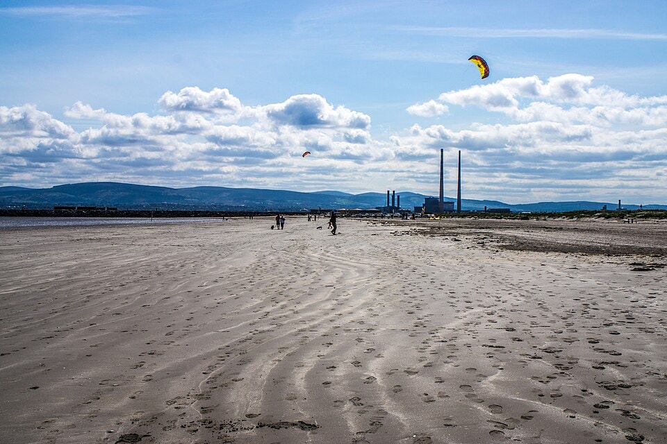 A kite flies over a vast sandy beach where several people are enjoying the open space near the water.