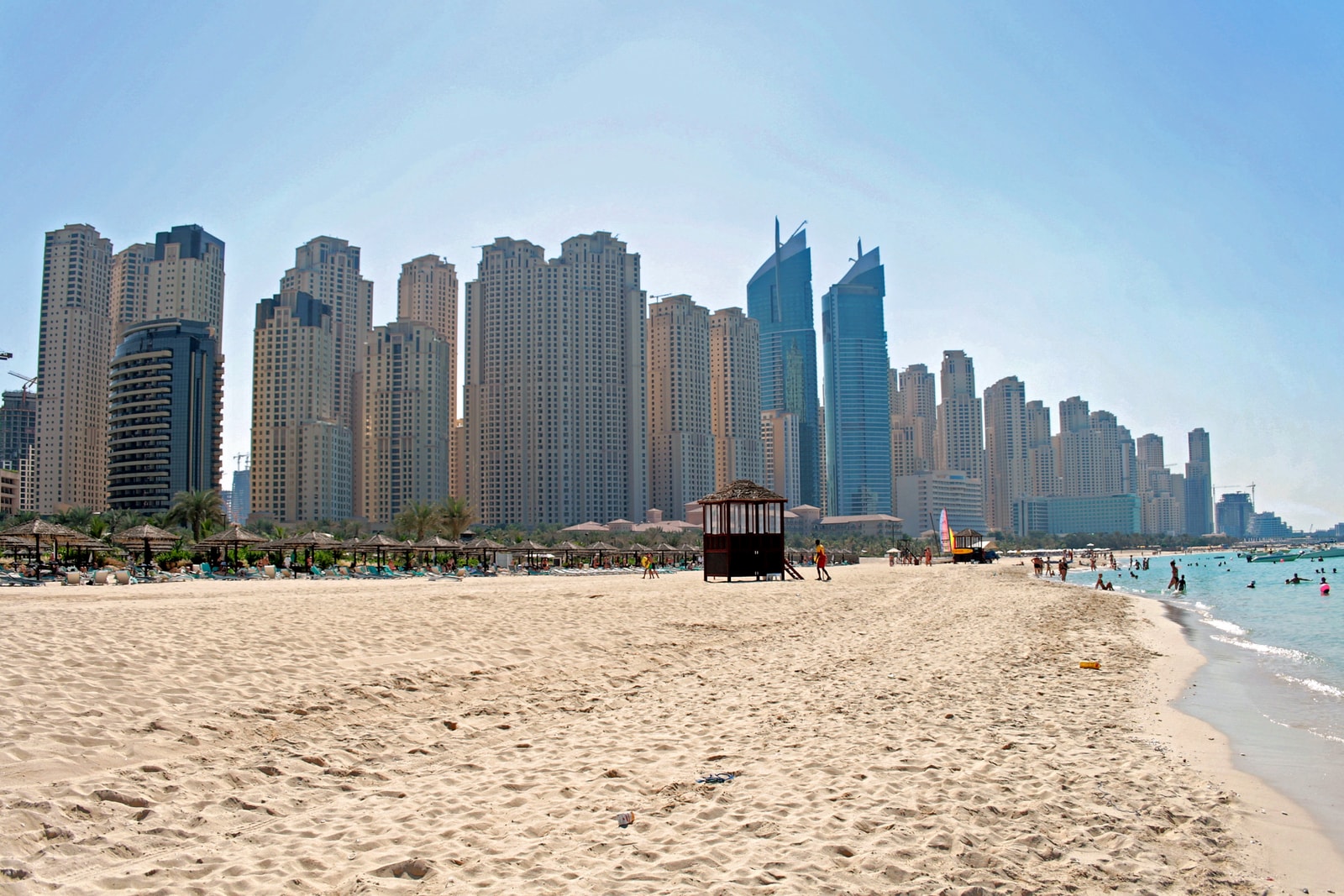 A broad view of a Dubai beach with a sandy foreground leading to the water, with the dense Marina skyline in the background.