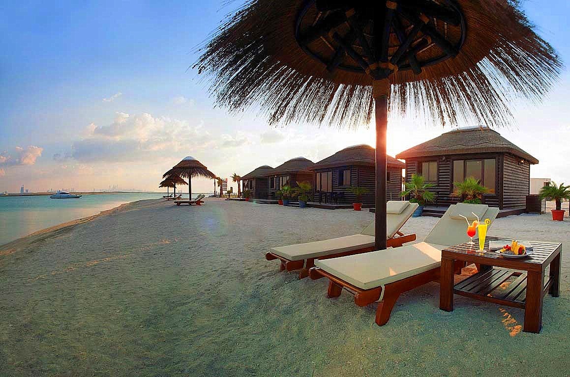 Beachfront cabanas and loungers are set up on a white sandy beach, overlooking calm turquoise water under a bright sky.