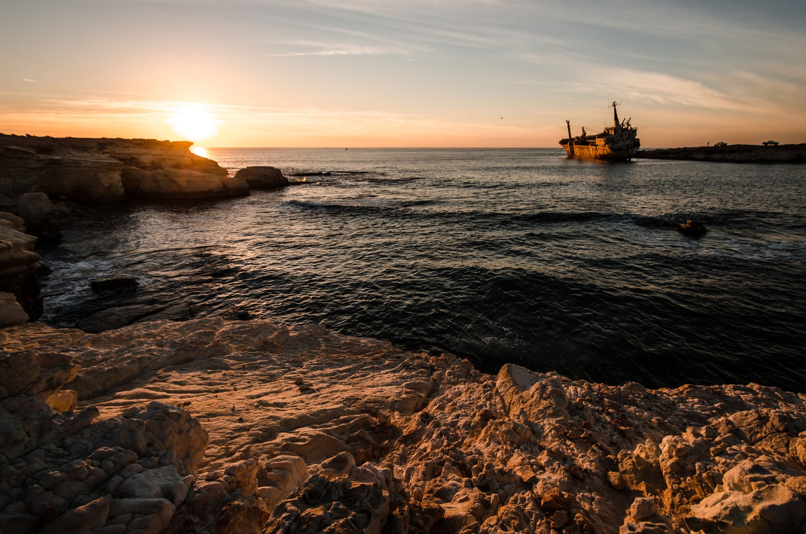 The silhouette of the Edro III shipwreck against a sunset sky, with a rocky foreground.