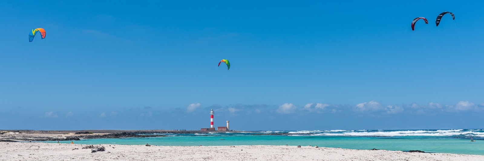 A panoramic shot of a beach with numerous kitesurfers on the water and in the air against a blue sky.