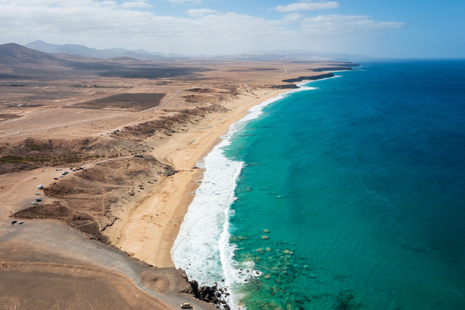 An aerial view of a wide, golden sand beach with turquoise water lapping at the shore and dark volcanic hills in the background.