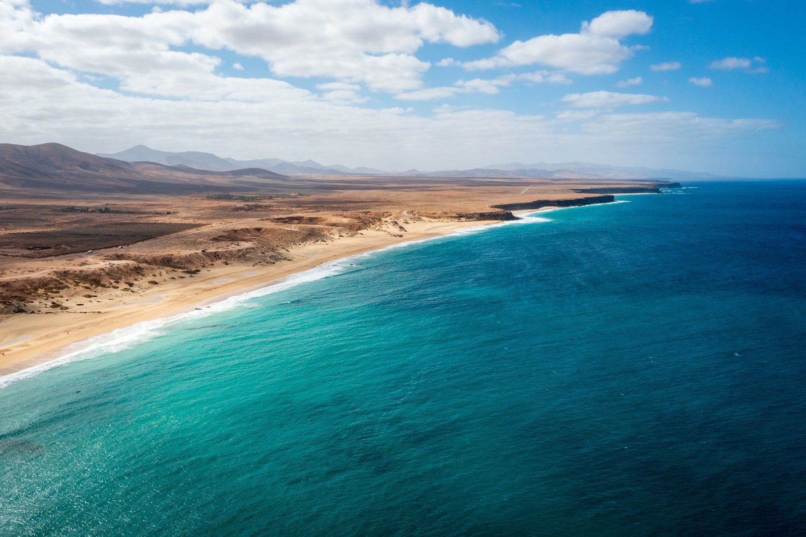 An aerial shot shows a wide, sandy beach on El Cotillo's west coast, with white surf crashing against the shore.