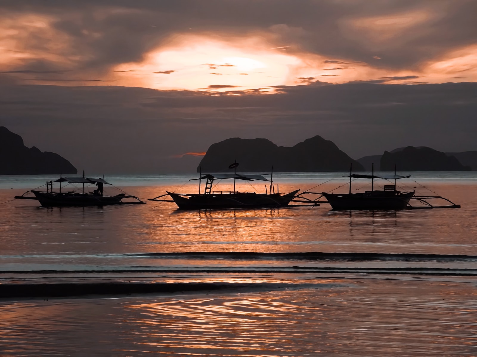 Several outrigger boats are silhouetted against a golden sunset sky in El Nido.