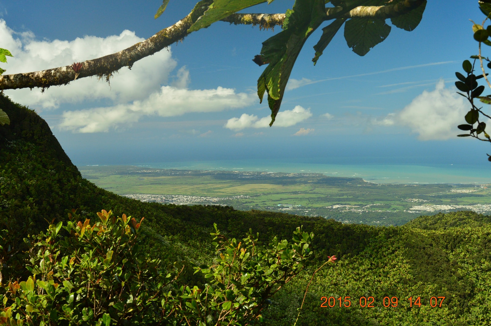A scenic view from a mountain peak looks out over a green coastal plain to the turquoise ocean.