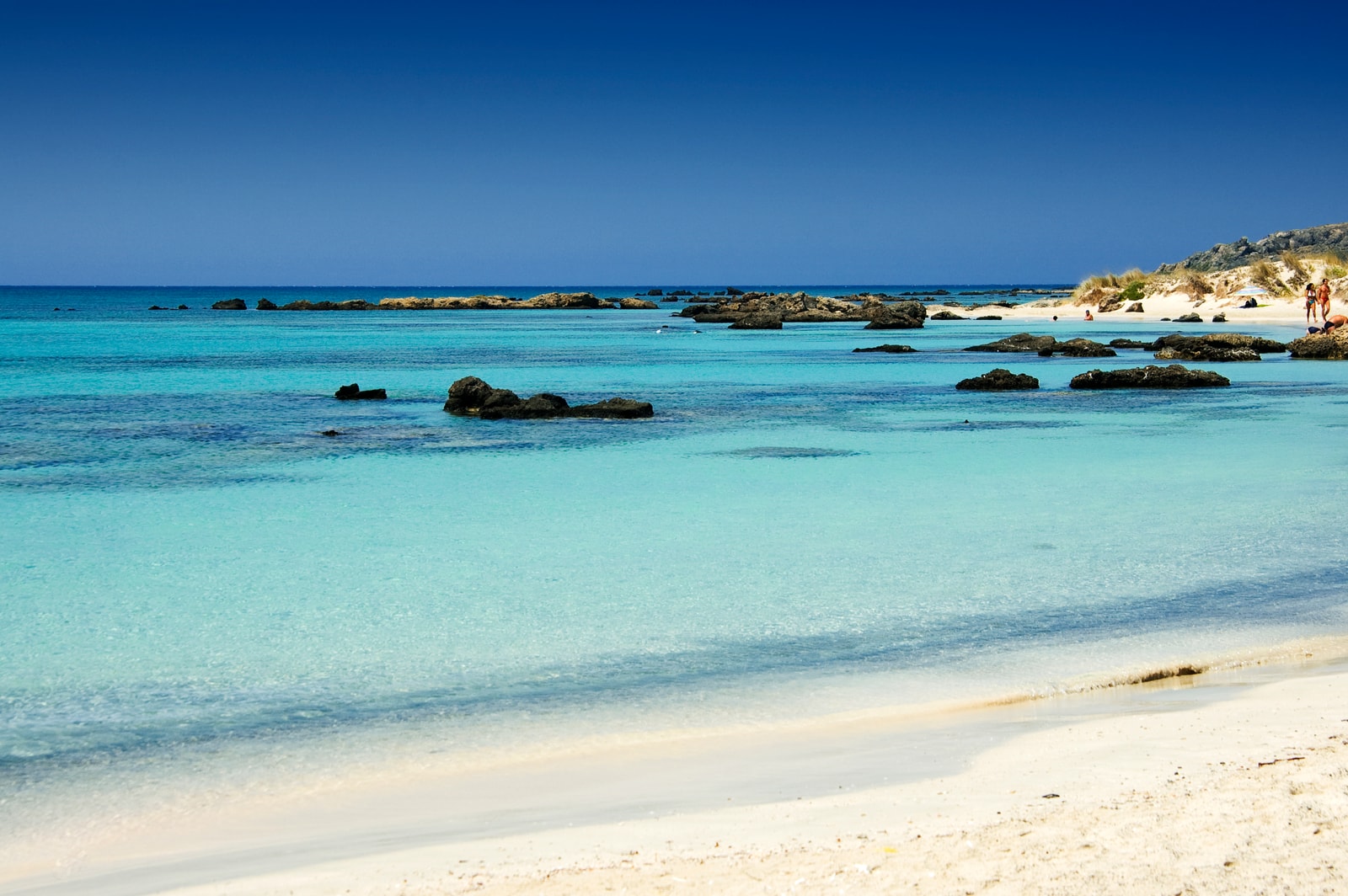 People wading through the shallow, calm waters of Elafonisi Beach under a clear sky.