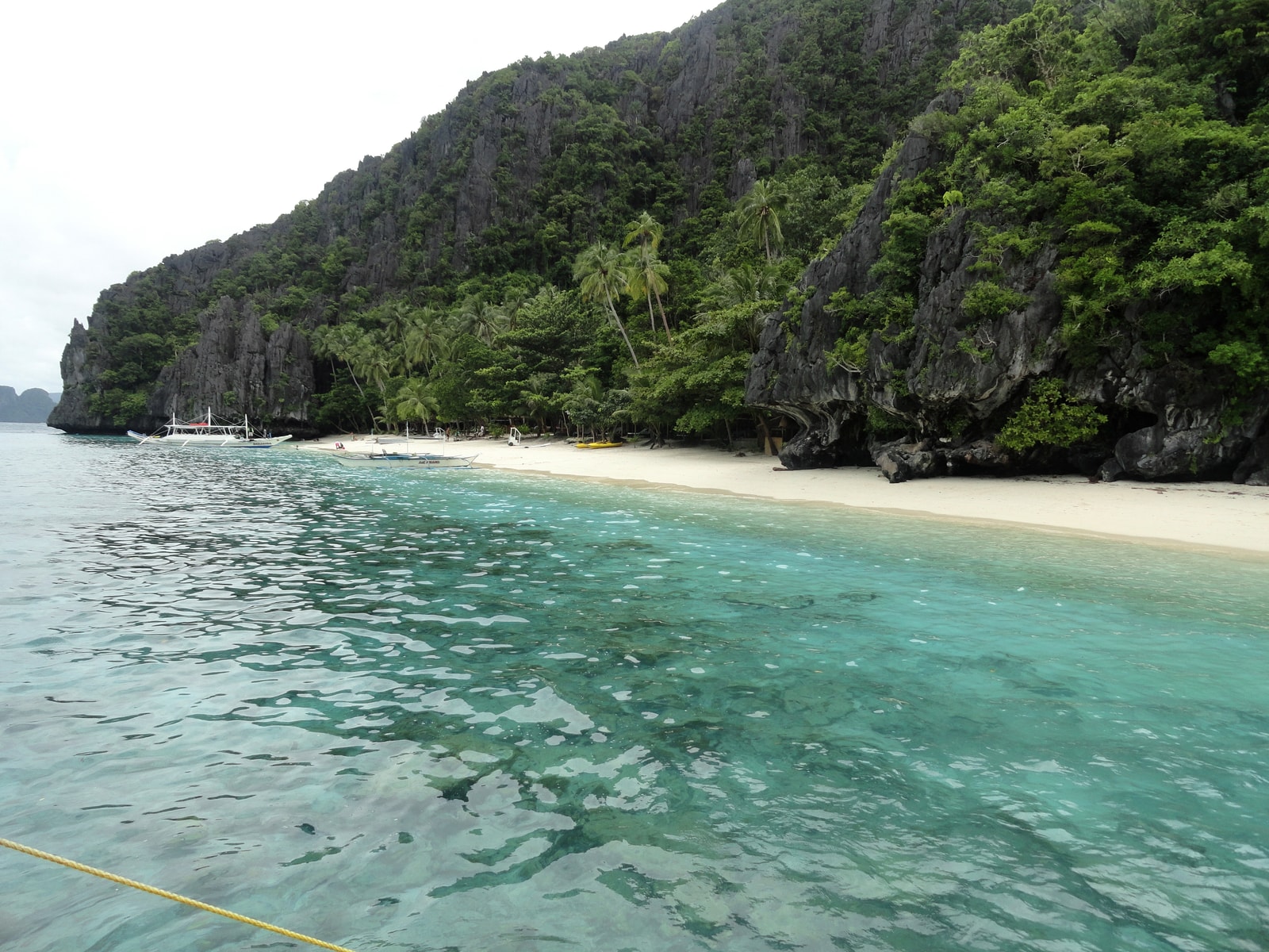 A view from the water shows the white sand beach of Entalula Island, with outrigger boats moored in the clear shallows.