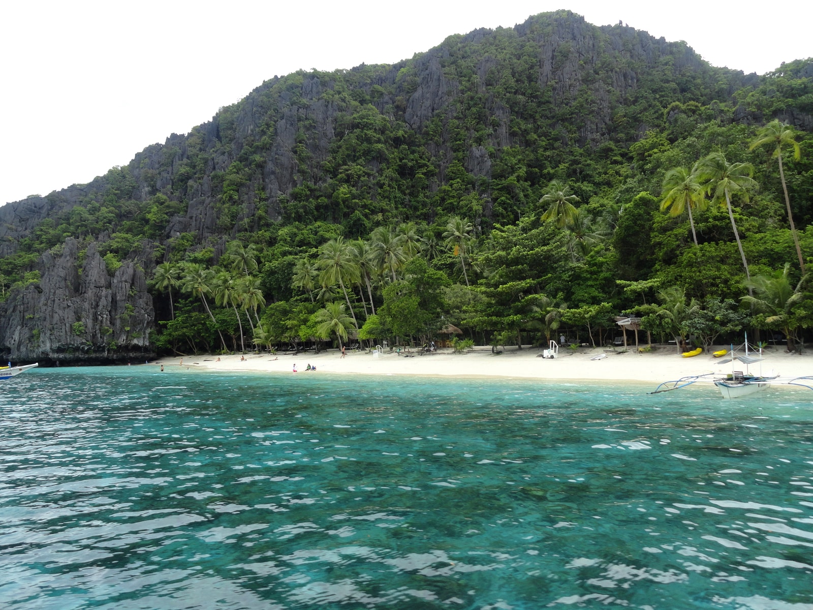 A broad view of Entalula Beach shows palm trees, white sand, bright blue water, and a towering karst backdrop.