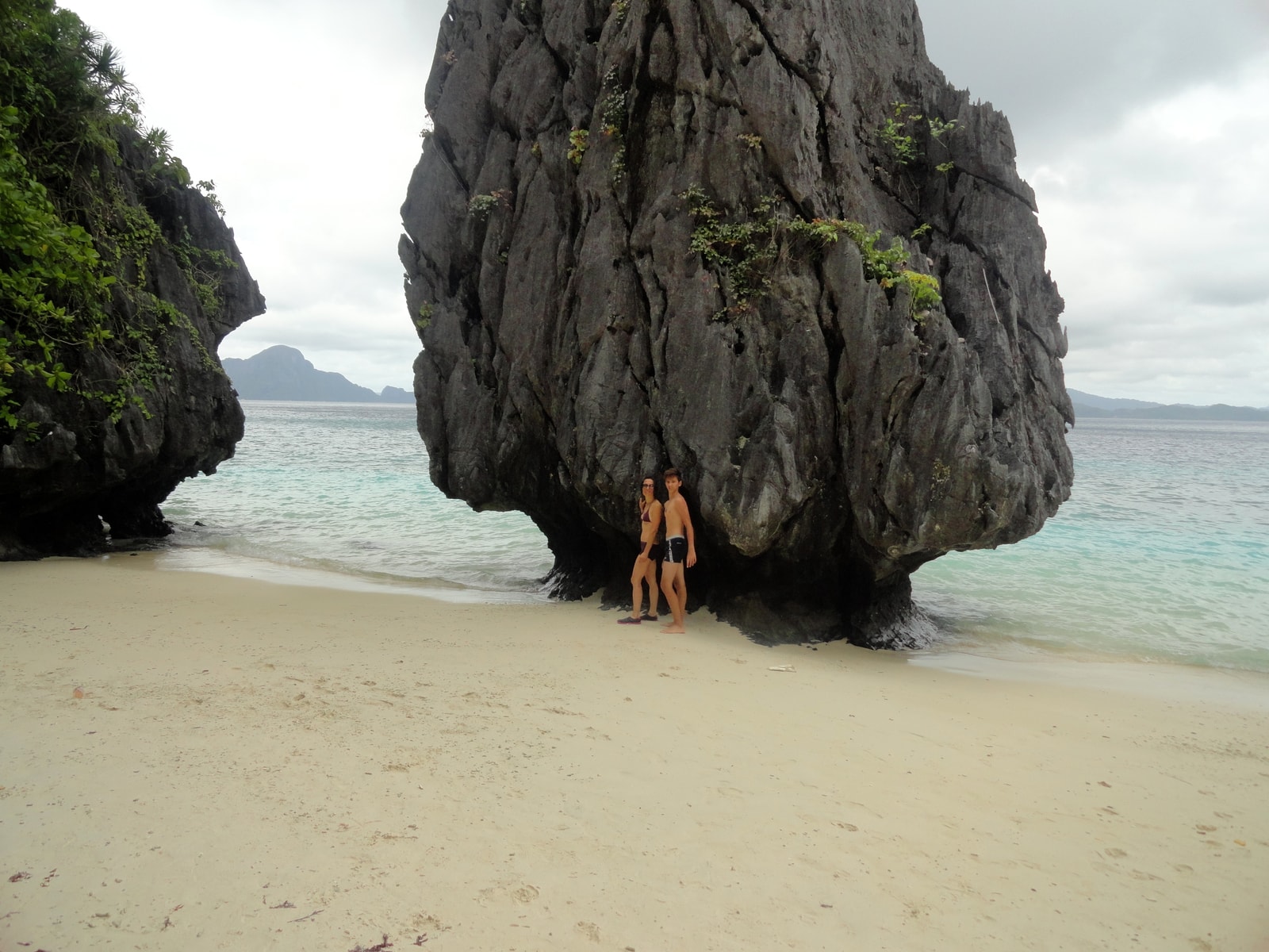 A dramatic overhanging limestone cliff shelters a small white sand beach where people are gathered.