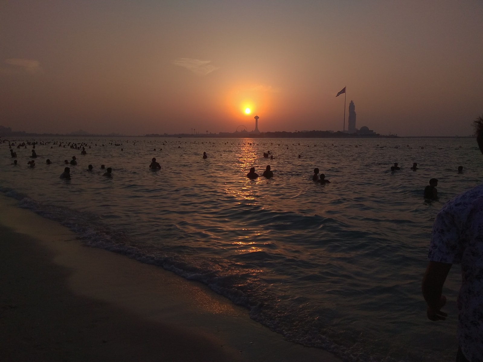 People wade and swim in the calm water of Corniche Beach during a warm evening, with the city lights in the background.