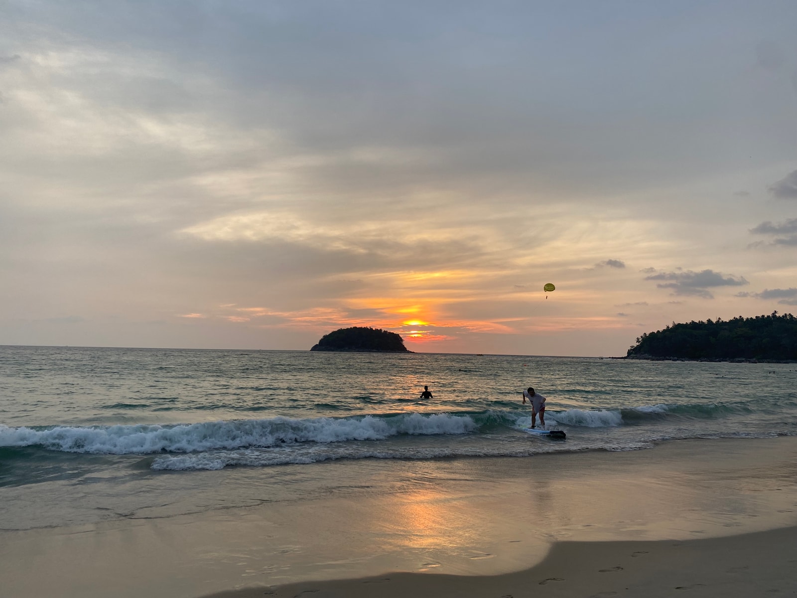 A warm sunset over Kata Beach, with silhouettes of the headland and clouds reflecting on the wet sand.