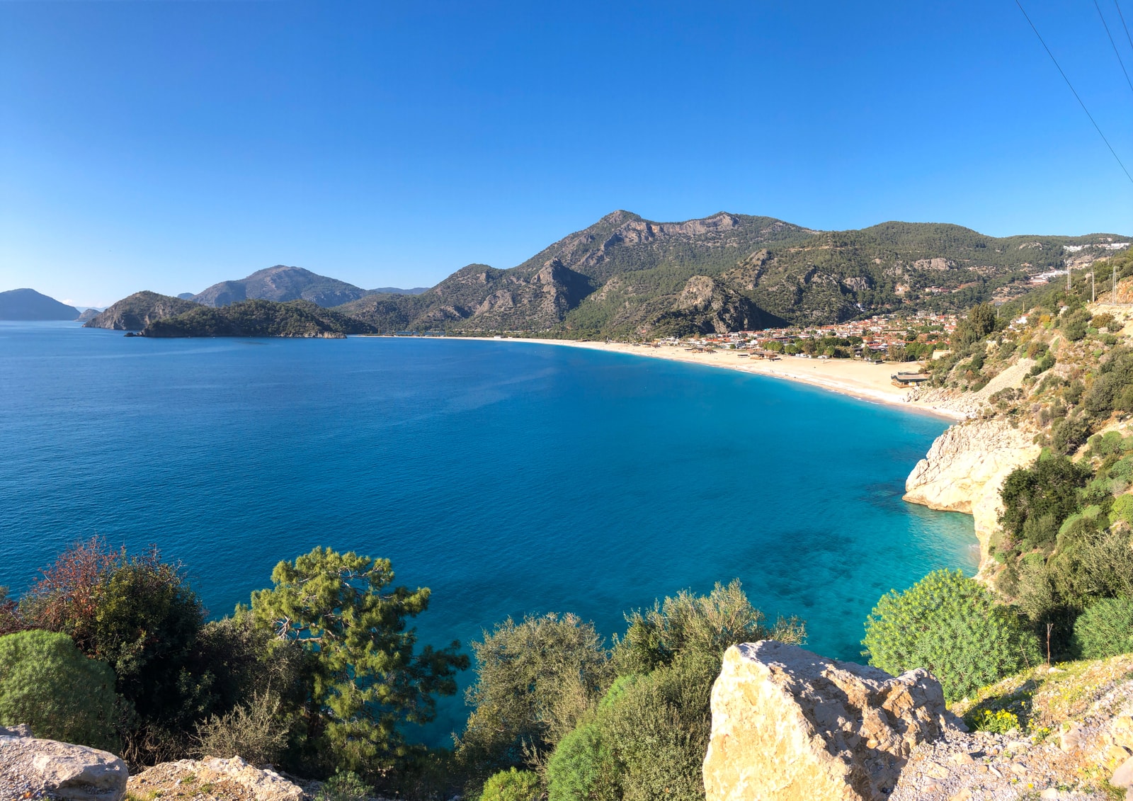 A wide panoramic view of Ölüdeniz bay, showing the curve of the beach and the green hills surrounding it.