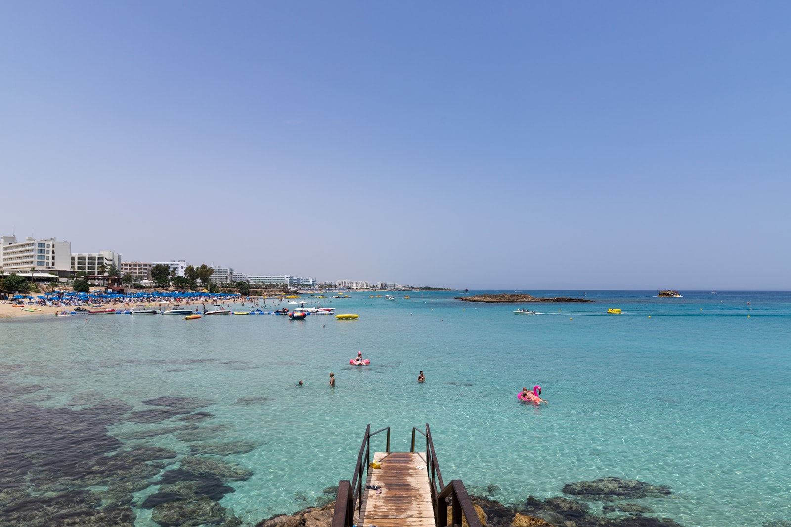 An aerial view of Fig Tree Bay, capturing the pier, numerous swimmers in the turquoise water, and boats moored nearby.