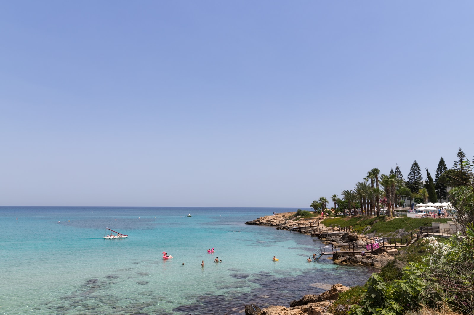 A view of a smaller Protaras beach, showing shallow turquoise water, a rocky point with greenery, and beachfront buildings.