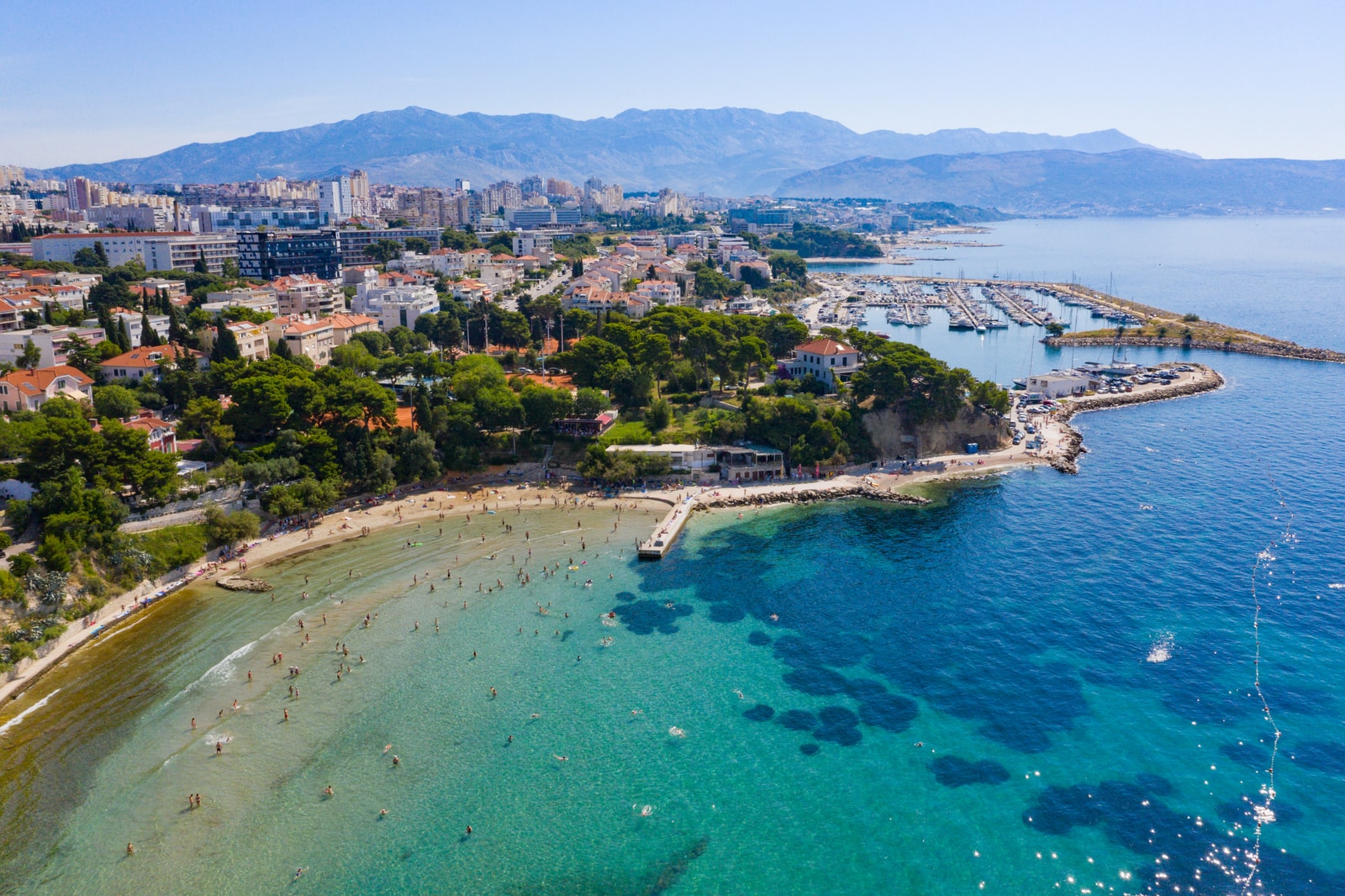 High-quality aerial view looking down at the sandy shore of Firule Beach and sailboats docked in Marina Zenta.