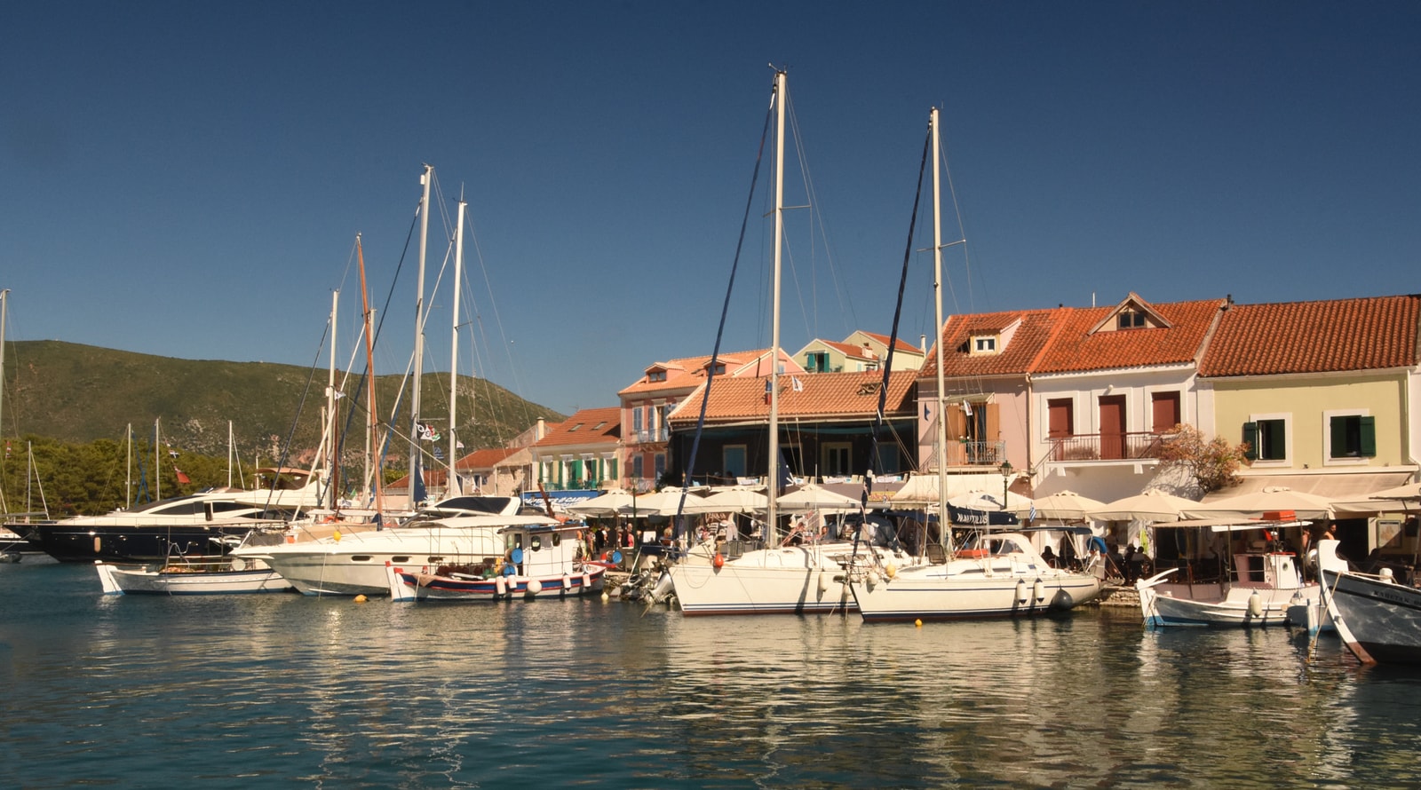 Luxury sailing yachts and boats moored in the bustling harbor of Fiskardo, with colorful waterfront buildings in the background.