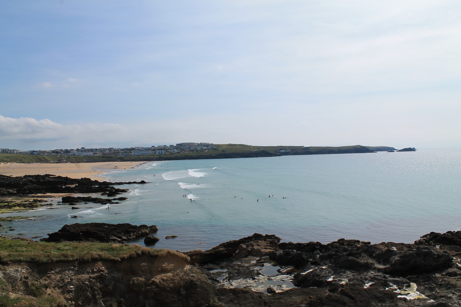 A panoramic view of Fistral Bay on a sunny day, showing surfers in the turquoise water and the town in the distance.