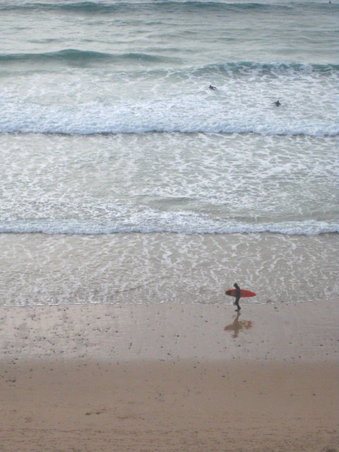 A lone surfer walks across the wet sand at sunset, carrying a red surfboard.