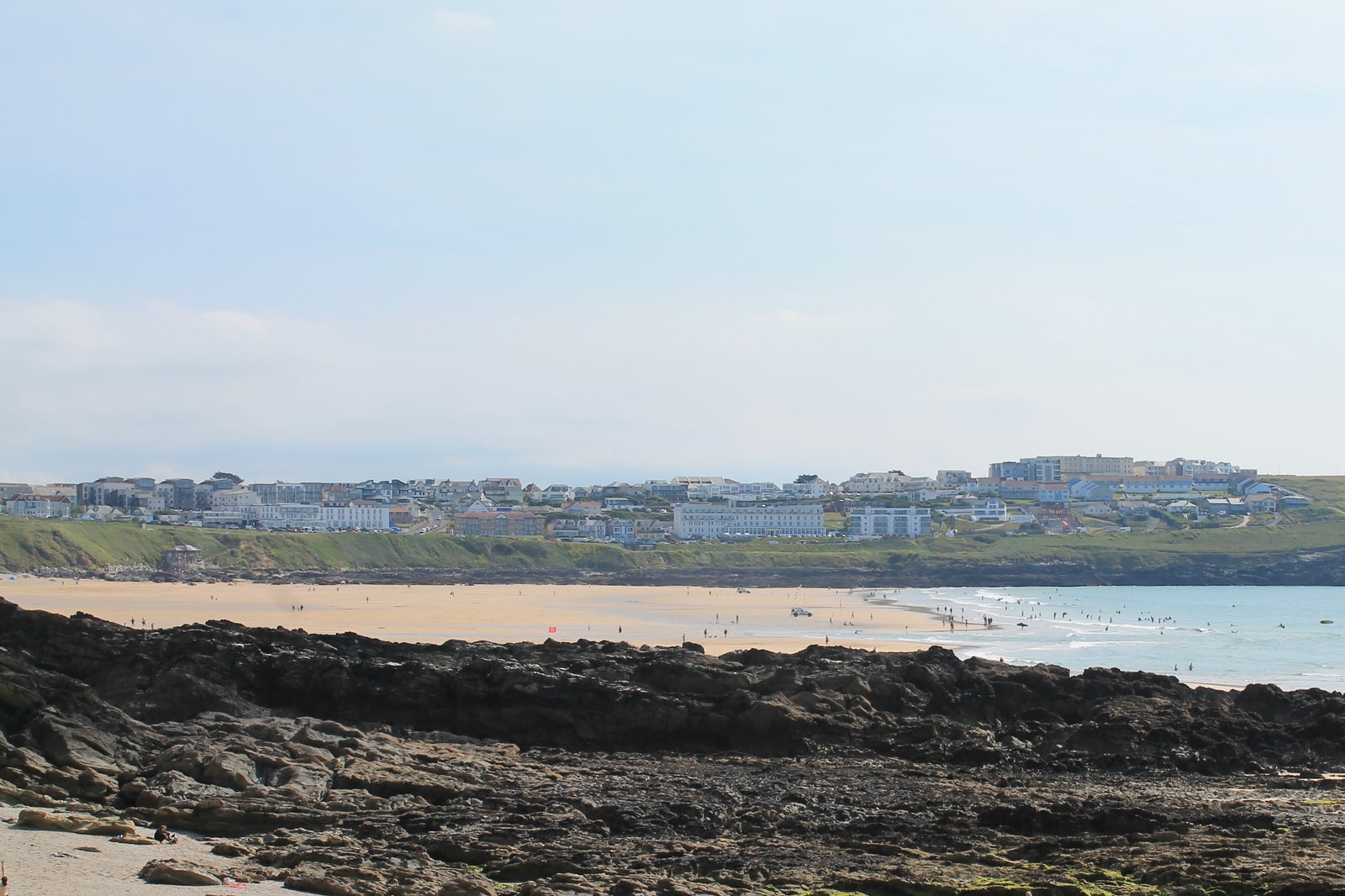A wide panoramic view of Fistral Beach showing the golden sand, dark shoreline rocks, and the town above.