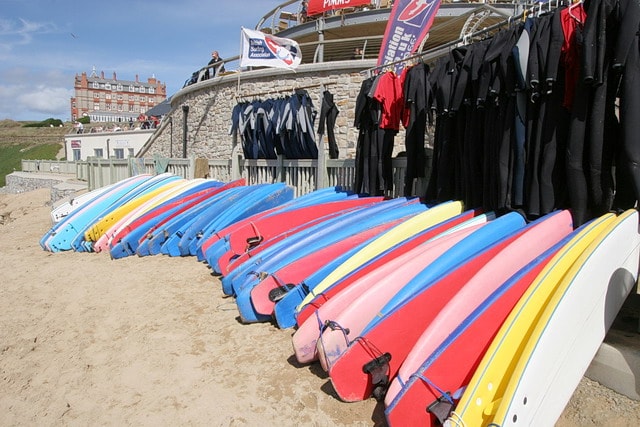 Colorful surfboards and wetsuits are lined up at a surf school on Fistral Beach, with the Headland Hotel in the background.