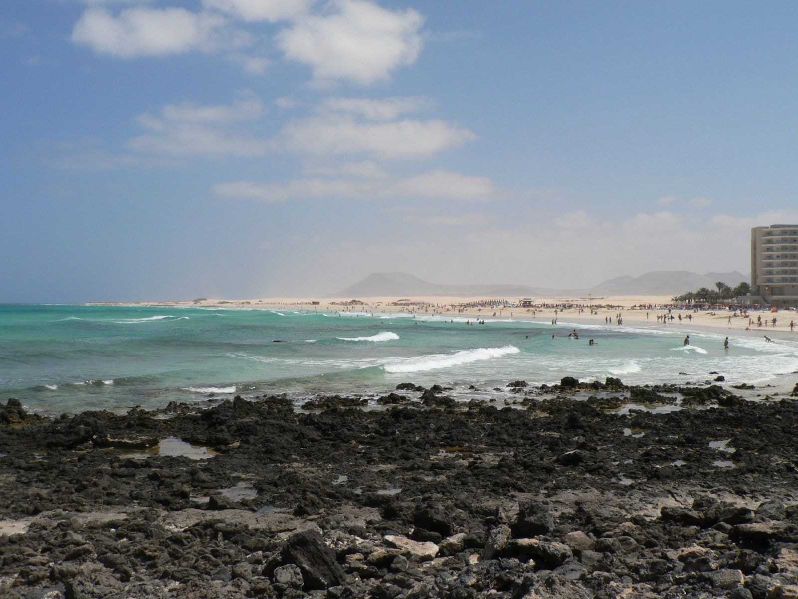 A sandy beach with dark volcanic rocks in the foreground, with turquoise water and the island of Lobos on the horizon.