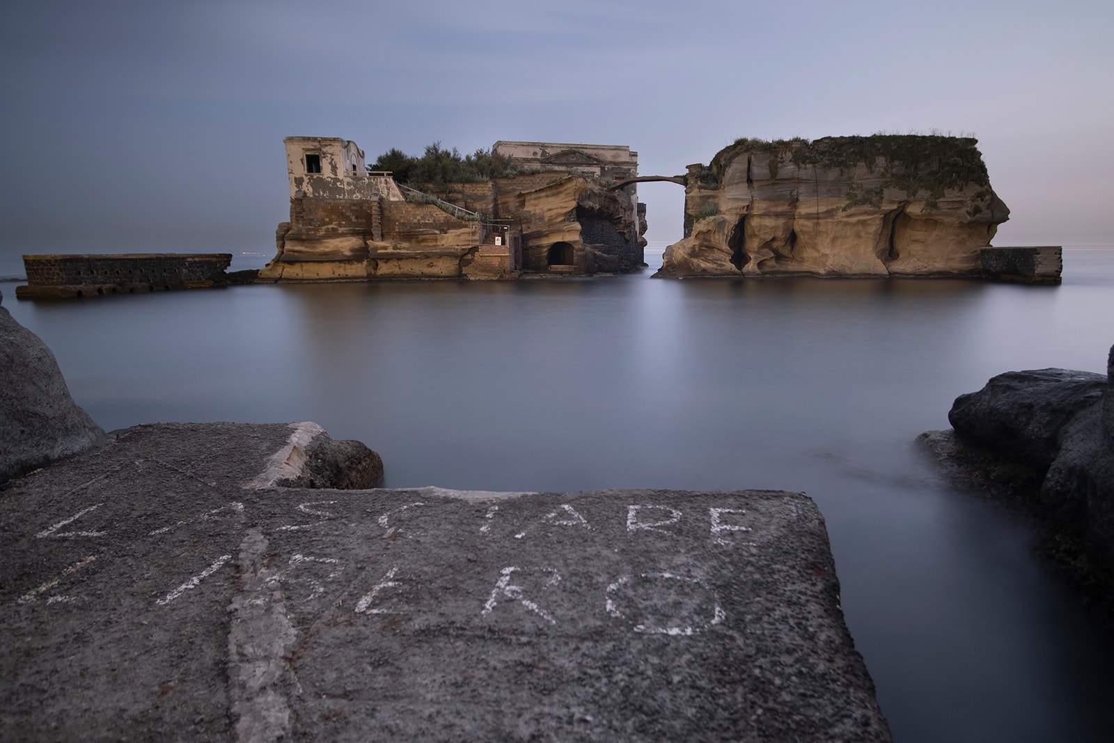 A long exposure shot of the two Gaiola islets at twilight, connected by a thin stone arch bridge, with silky smooth water surrounding them.