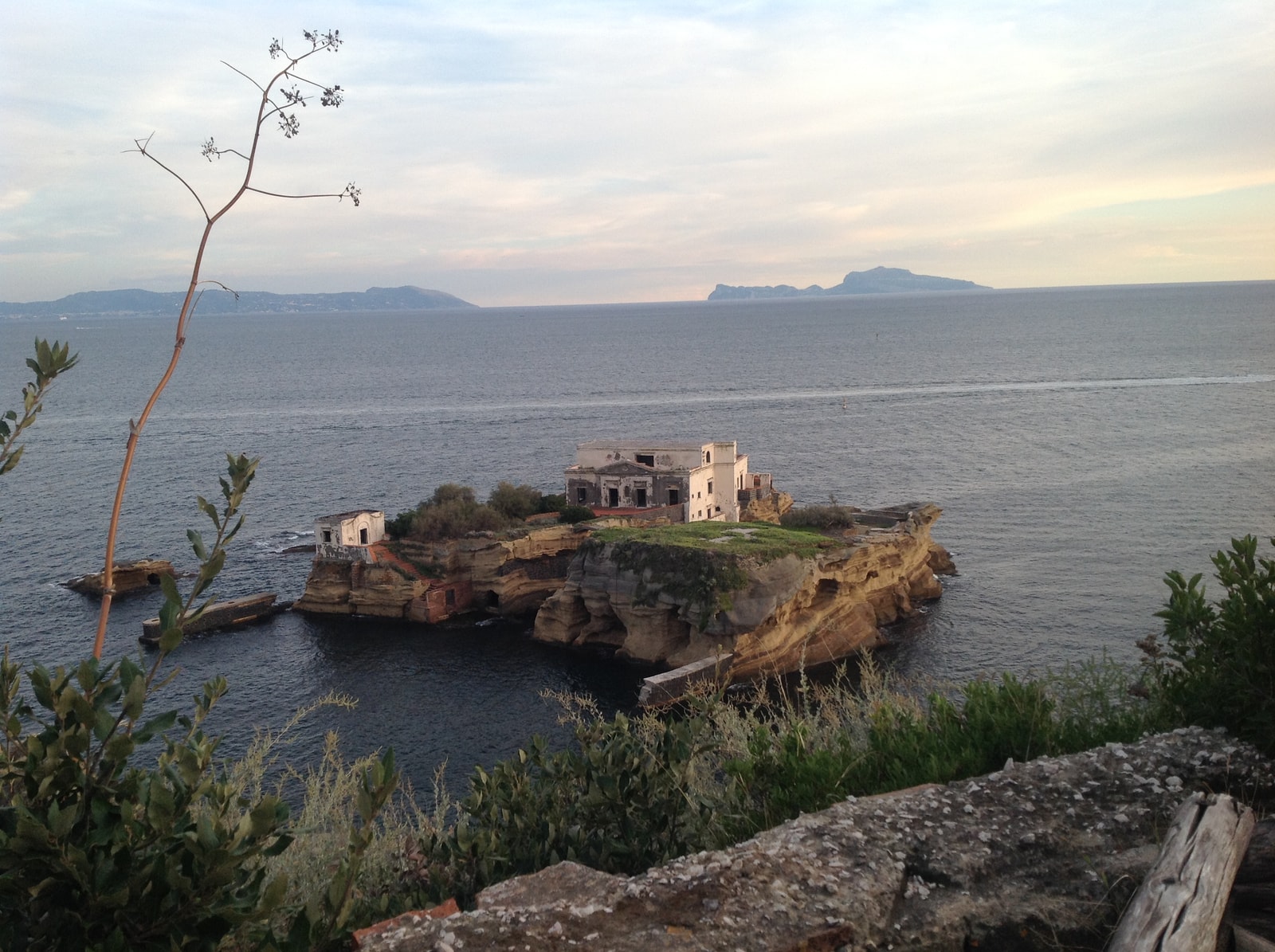 An elevated view of one of the Gaiola islets, showing the abandoned white villa surrounded by lush greenery and the clear blue waters of the protected park.