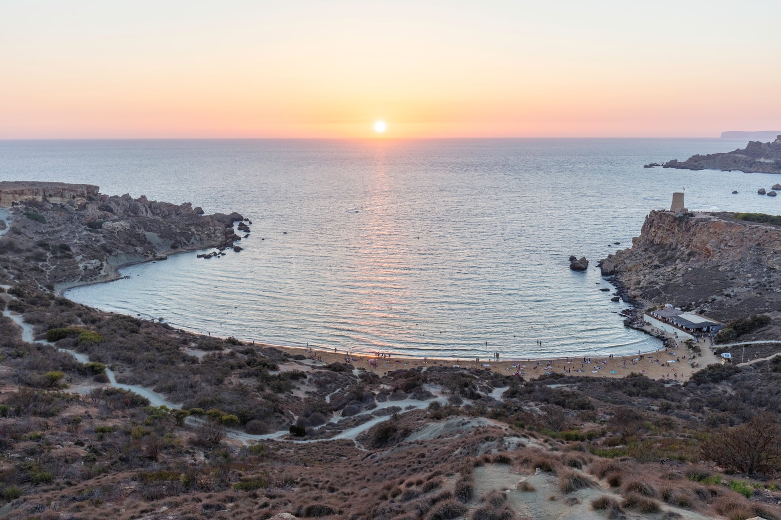 A stunning high-angle view of the sandy Għajn Tuffieħa Bay (Riviera Beach) glowing at sunset, with a historic watchtower on the cliffs.