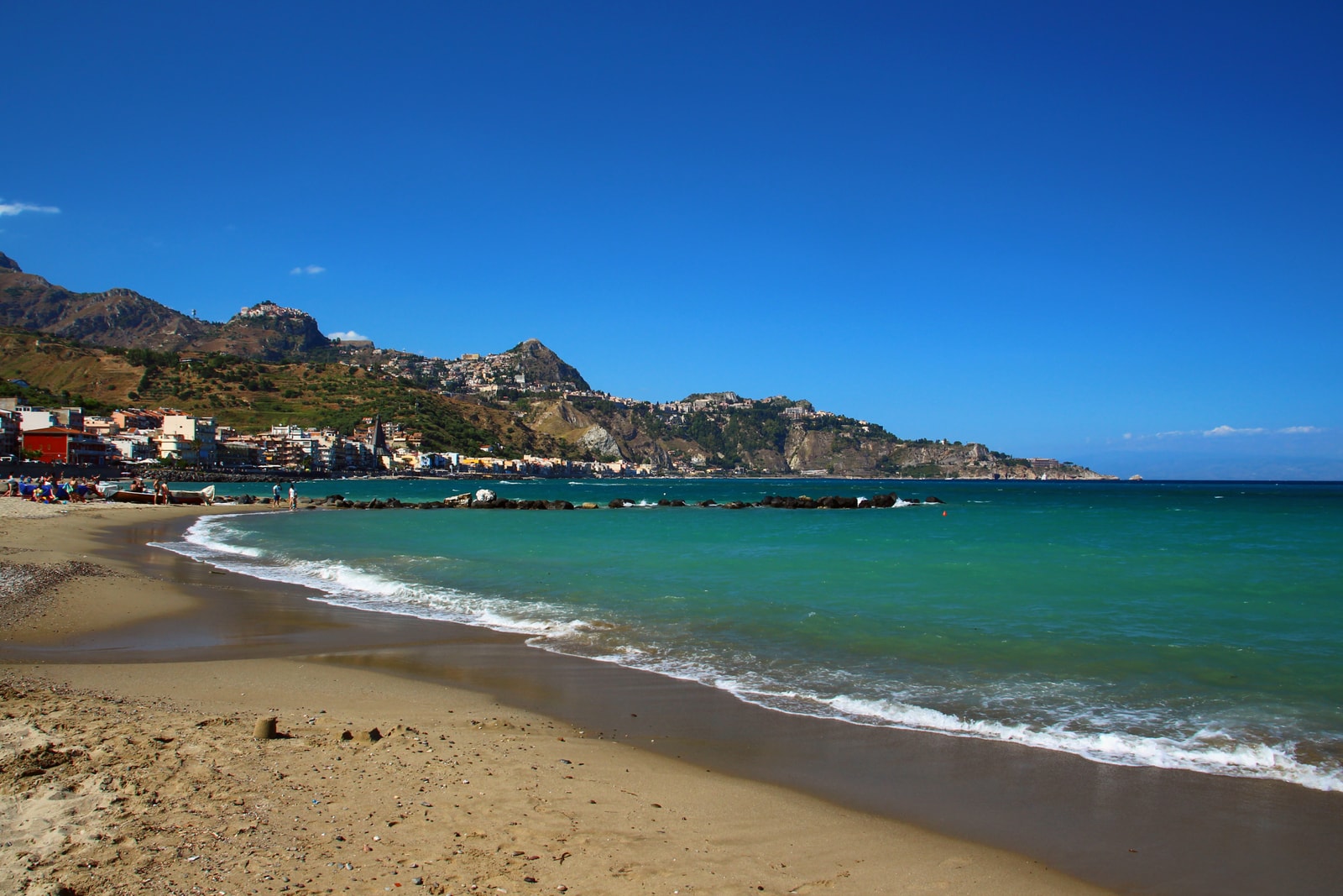 Broad sandy resort beach of Giardini Naxos with shallow turquoise waters and surrounding hills on a clear summer day.
