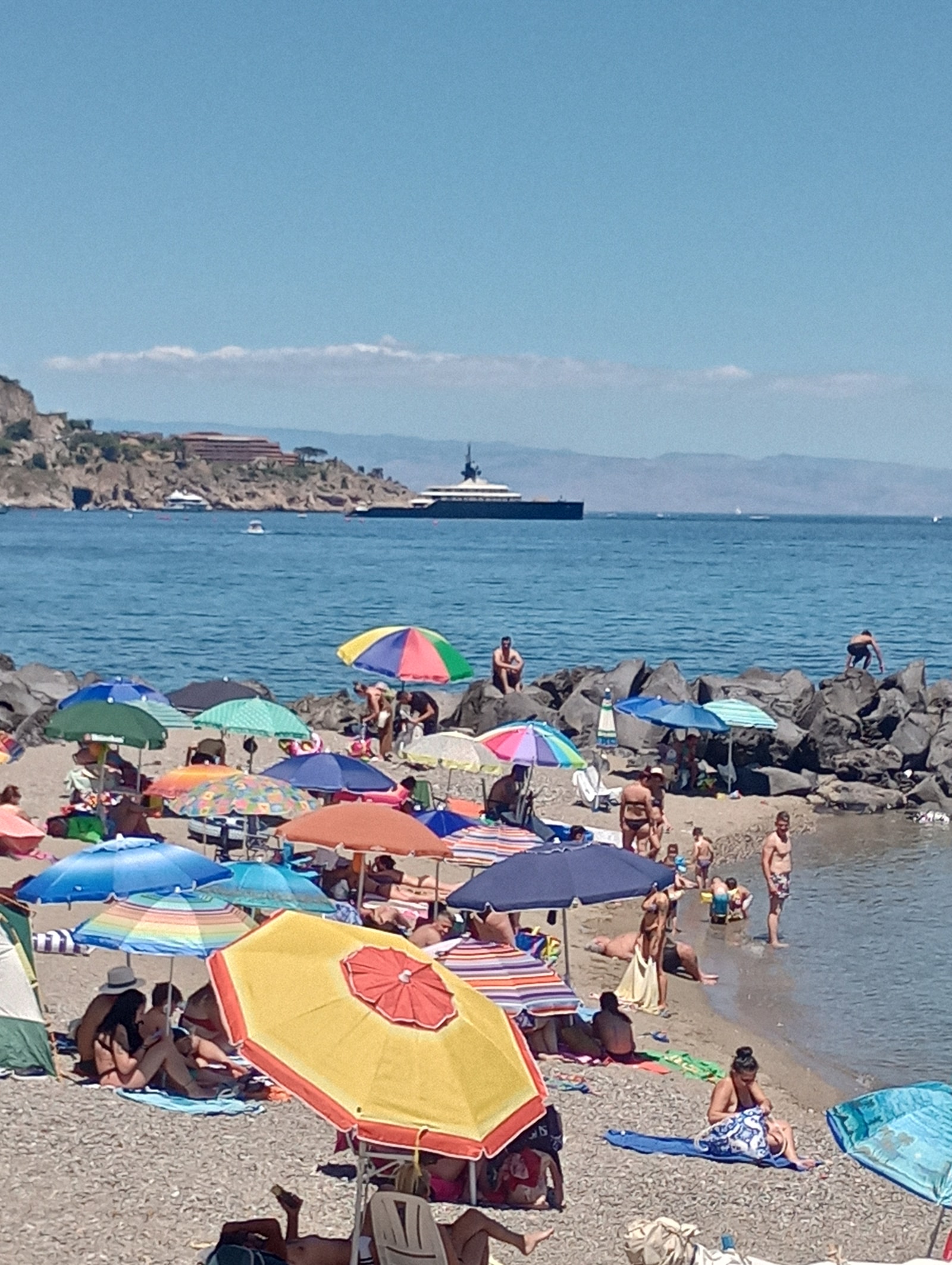A busy summer day at Giardini Naxos, with rows of colorful beach umbrellas and a luxury superyacht anchored in the bay.
