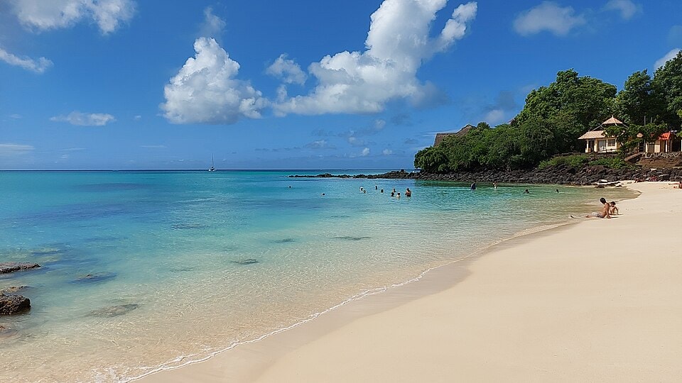 A beautiful shot of La Cuvette Beach in Grand Baie, with clear turquoise water, white sand, and lush green trees.