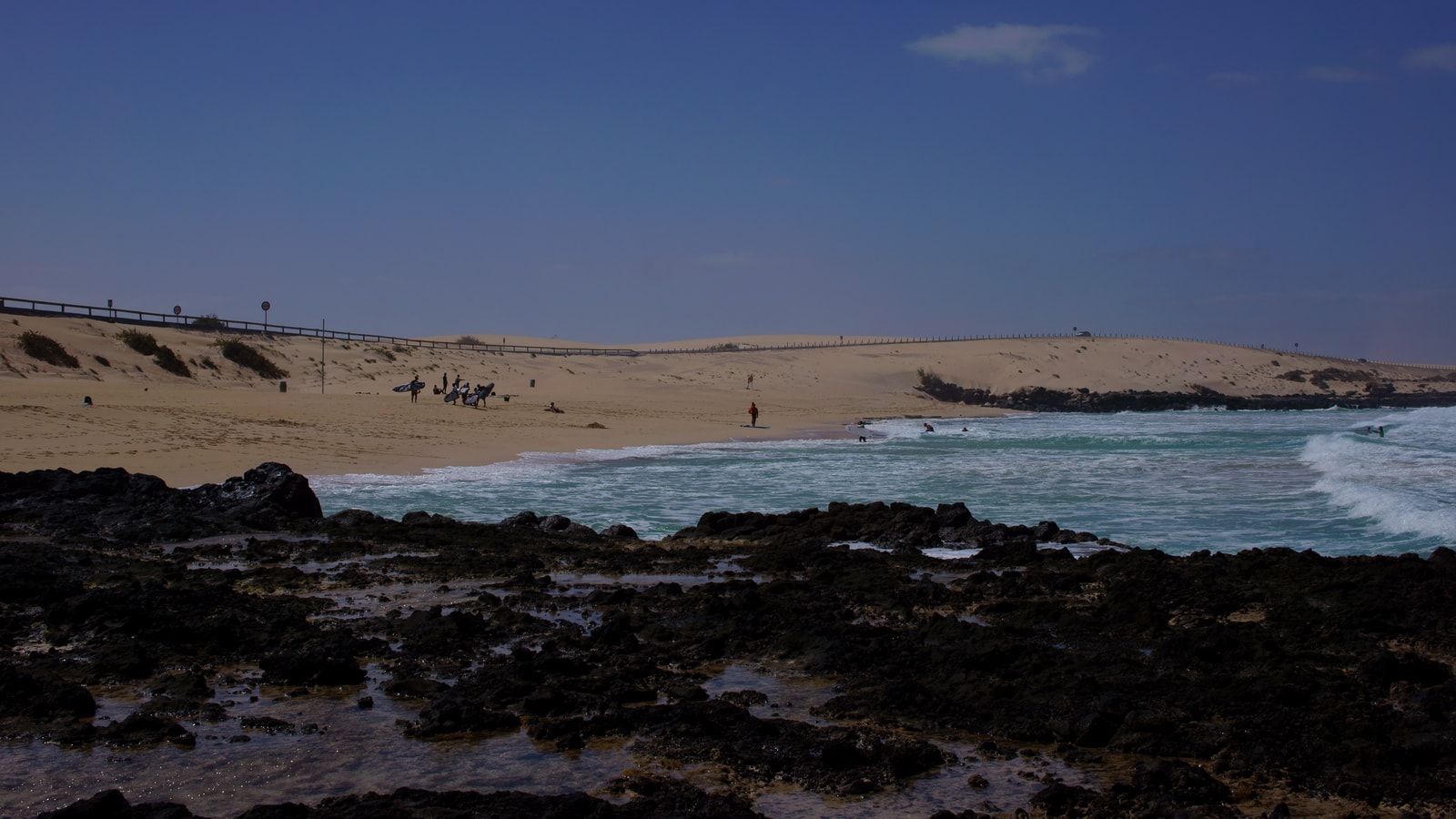 A wooden walkway crosses white sand dunes, leading towards a sparkling turquoise sea with beachgoers in the distance.