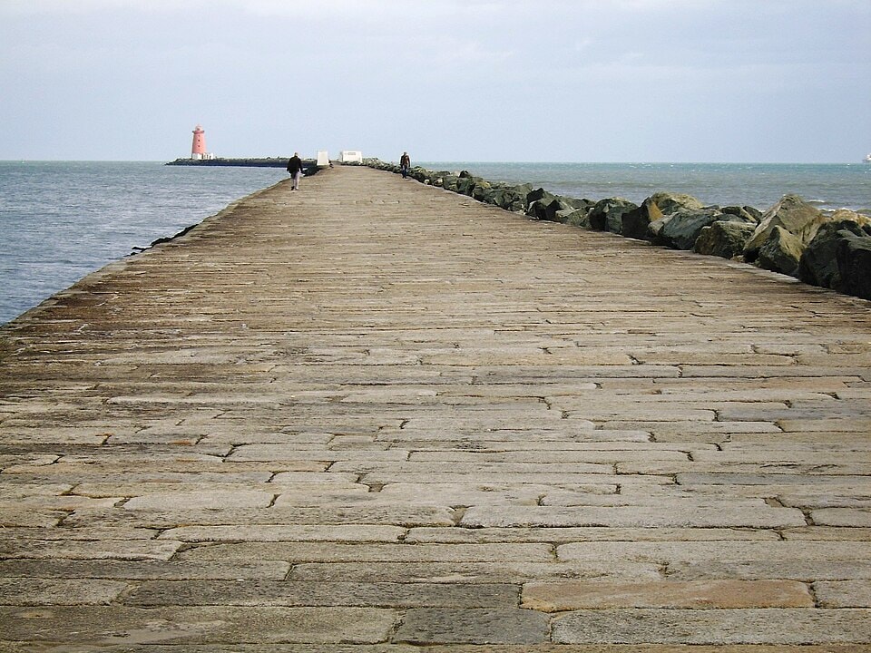 A long stone breakwater stretches far out into the water toward a red lighthouse.