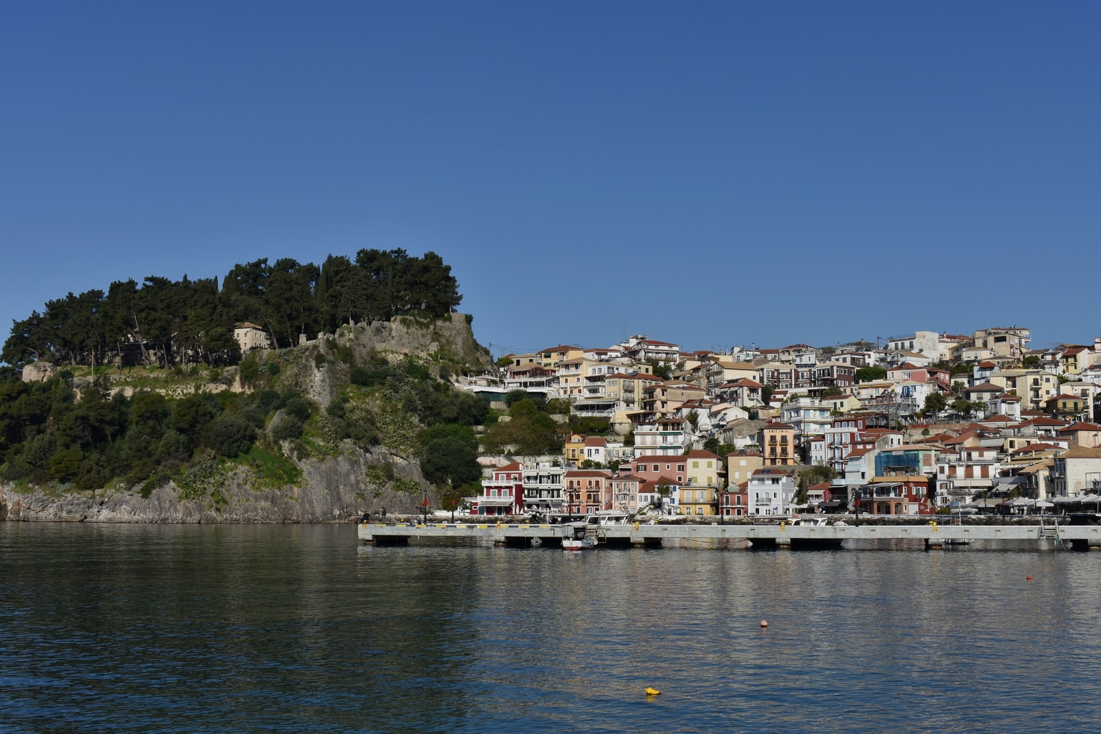 A high-angle view of Parga town, showing colorful buildings climbing up a hill next to the sea and the Venetian castle on a promontory.
