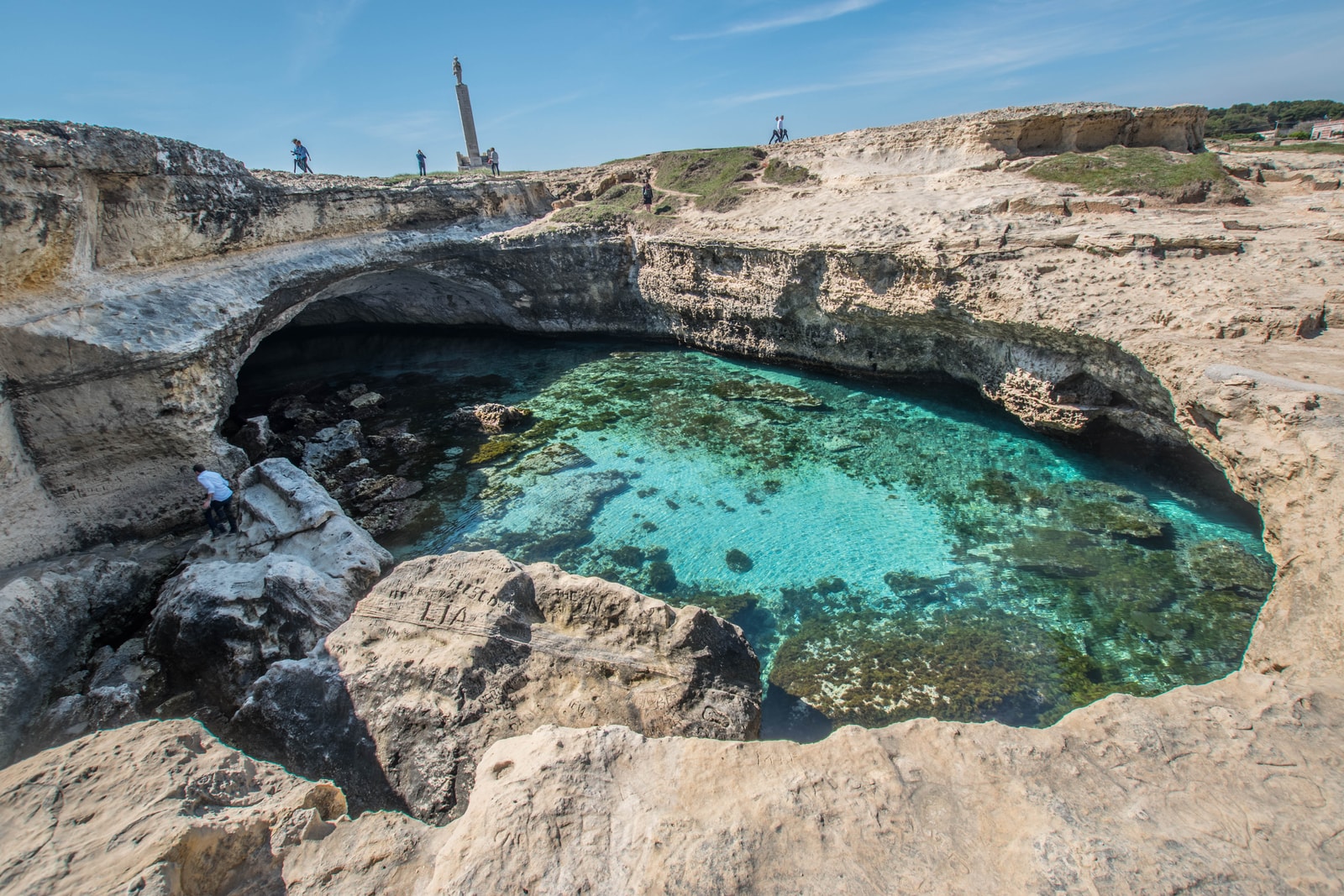 A high-angle view of the Grotta della Poesia, a circular natural swimming pool formed by a collapsed cave, with deep turquoise water.