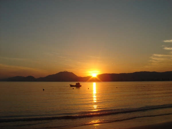 A calm bay in Guarujá at sunset, with boats on the water and mountains in the distance under a softly lit sky.