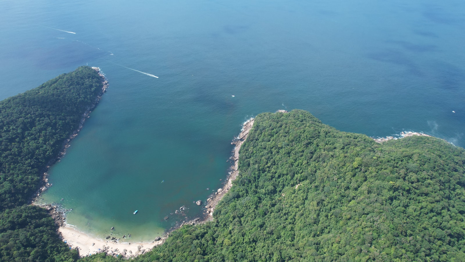 An aerial view of a secluded turquoise cove in Guarujá, surrounded by dense green forest and rocky headlands.