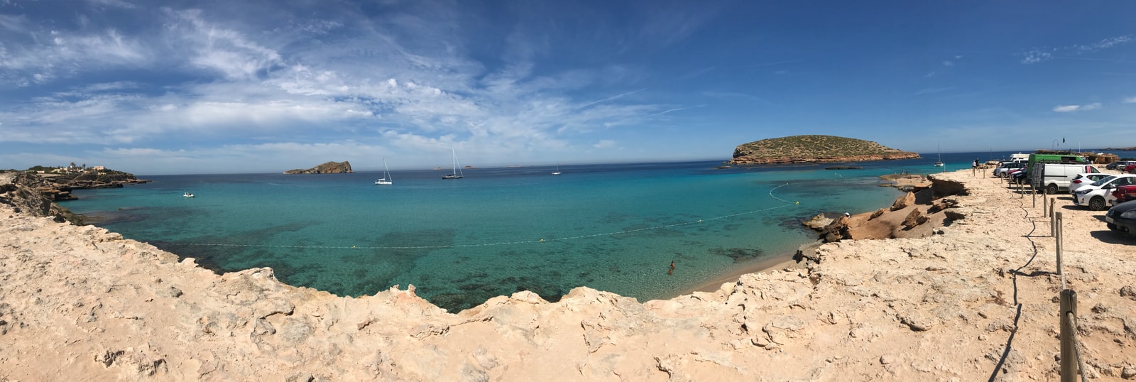 A comprehensive, wide panoramic view of Cala Comte showing parked campervans and s'Illa des Bosc in the distance.