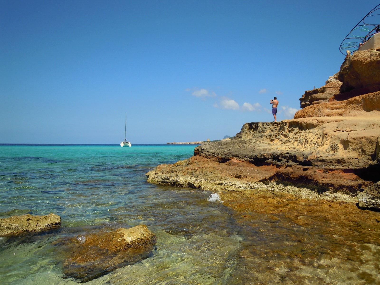 The rugged, layered orange and red sandstone cliffs of Cala Conta meeting the shallow, transparent turquoise water of the cove.