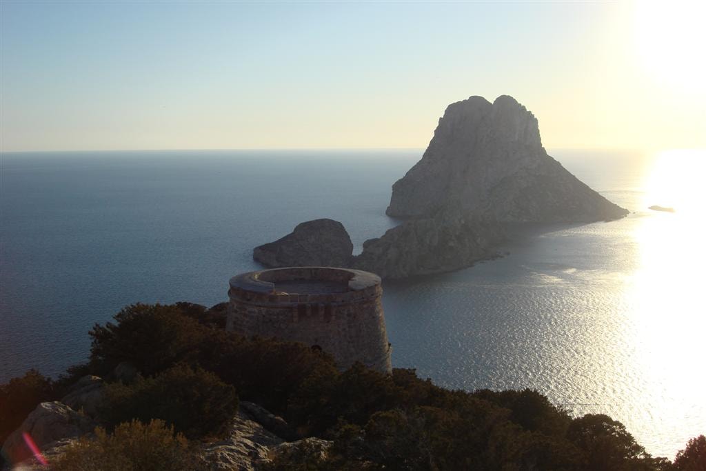The iconic Es Vedrà monolith rising from the deep blue sea, viewed from a high cliffside watchtower.