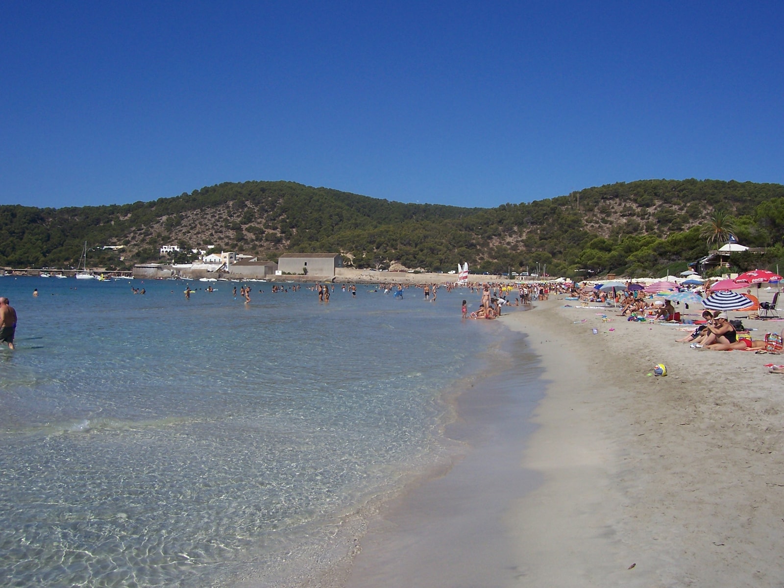 A bustling, sunny day on the sand of Ses Salines beach with colorful beach umbrellas and active beachgoers.